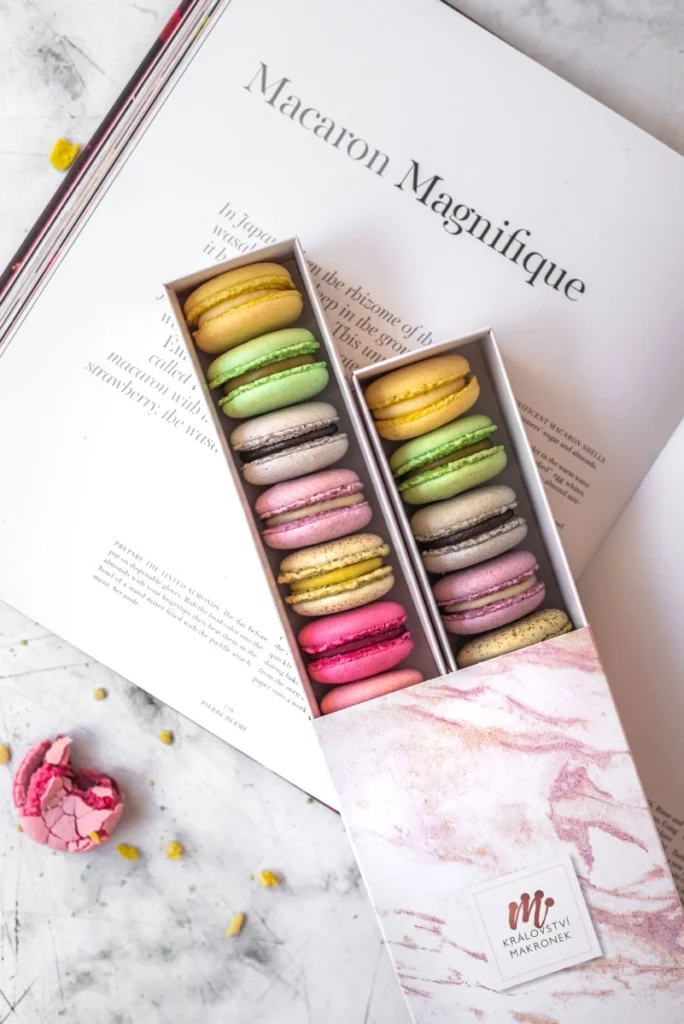 Elegantní krabička s barevnými makronkami v pastelových odstínech, naaranžovaná na otevřené knize s nápisem ‚Macaron Magnifique‘ na mramorovém stole.